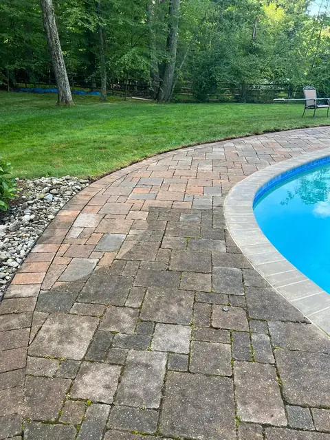 Paver patio beside a pool, darkened with moss and weeds